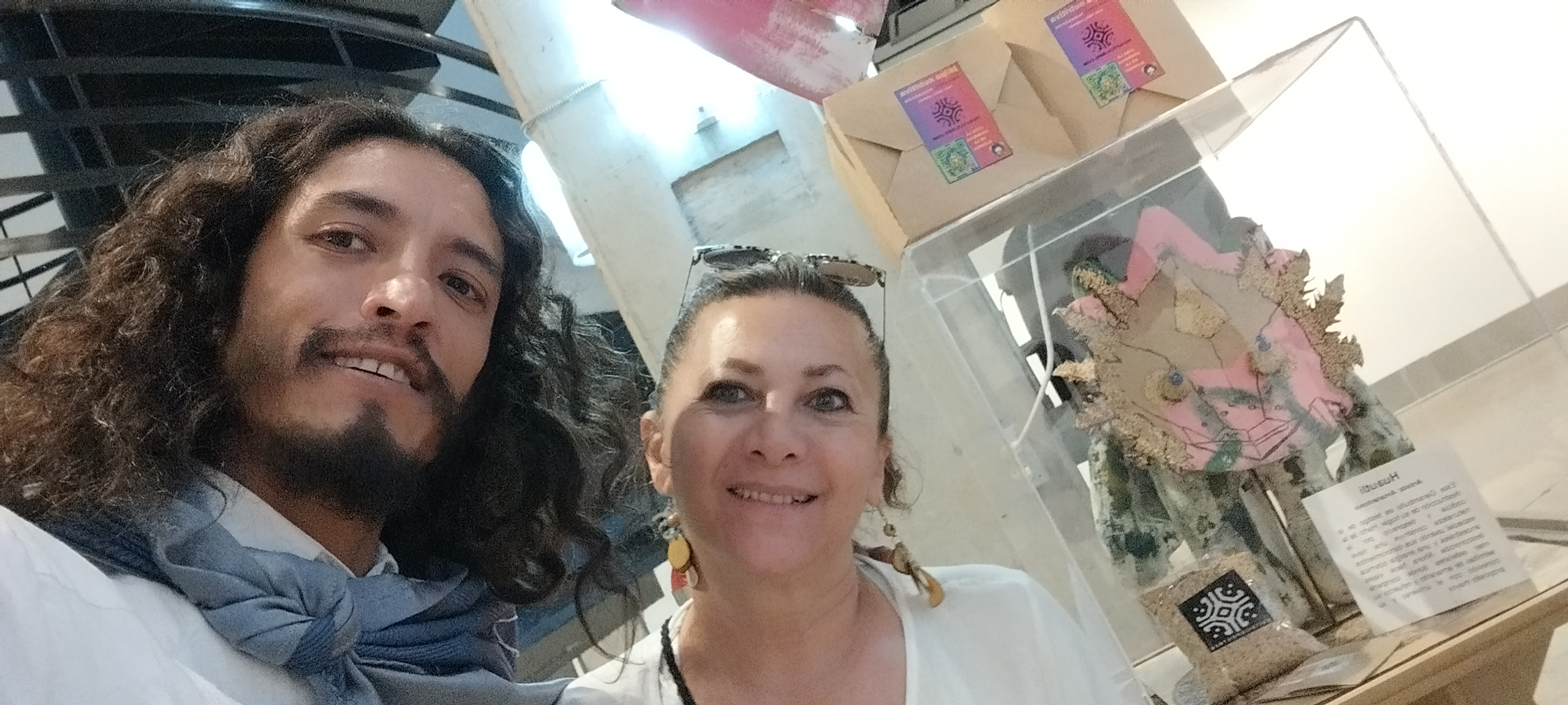Amarantonio y una mujer sonriendo frente a una exhibición de arte con una escultura y productos de amaranto.